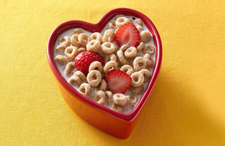 a full bowl of cheerios and milk, the bowl is in a heart shape and there are 3 halved strawberries in the bowl floating in the milk with the cereal