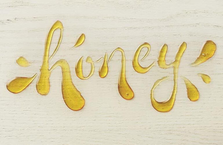 Le mot honey écrit avec le condiment miel sur une planche de bois, il y a des touches et des traits de miel des deux côtés du mot pour l'accentuer.