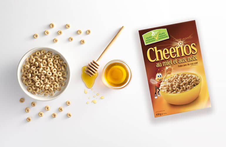 Un bol de céréales Cheerios, un bol de miel avec une cuillère et une boîte de Cheerios au miel et aux noix posés en ligne sur un fond blanc.