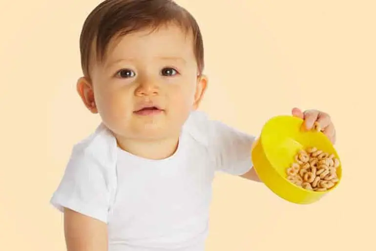 A smiling baby holding a plastic bowl in one hand at a precarious angle. In the bowl are cheerios which are almost falling out due to the angle