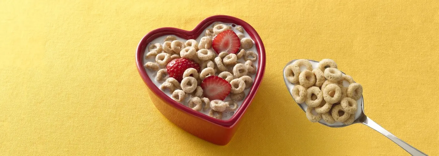 a full bowl of cheerios and milk, the bowl is in a heart shape and there are 3 halved strawberries in the bowl floating in the milk with the cereal