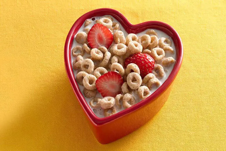 a full bowl of cheerios and milk, the bowl is in a heart shape and there are 3 halved strawberries in the bowl floating in the milk with the cereal