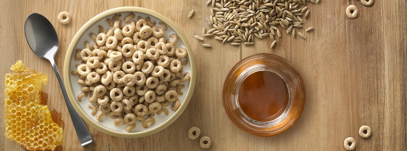 A honeycomb, a bowl of cheerios, a spoon, a jar of honey, and oats on a wooden table