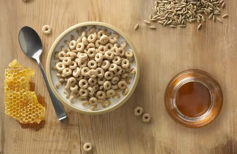 A honeycomb, a bowl of cheerios, a spoon, a jar of honey, and oats on a wooden table